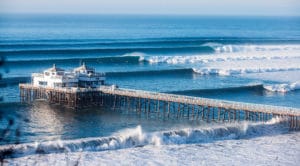 the Malibu pier surrounded by a bright blue ocean stacked with big waves breaking into foamy white wash