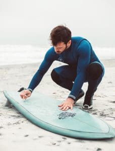 Man in a wetsuit waxing his surfboard.